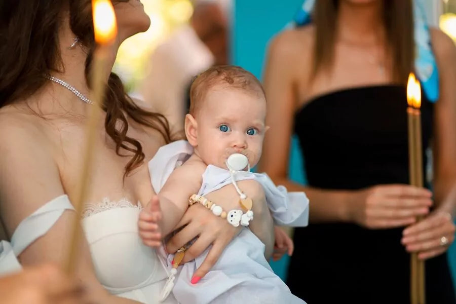 Bébé aux yeux bleus avec une sucette dans les bras de sa maman lors d’un baptême, entourée de bougies